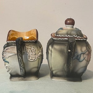 Op de afbeelding: Een vintage keramische suiker- en roomset met een drakenontwerp. De set is geschilderd in tinten grijs, wit, blauw en bruin. De roomkan heeft een bruin deksel en een gegolfde rand.