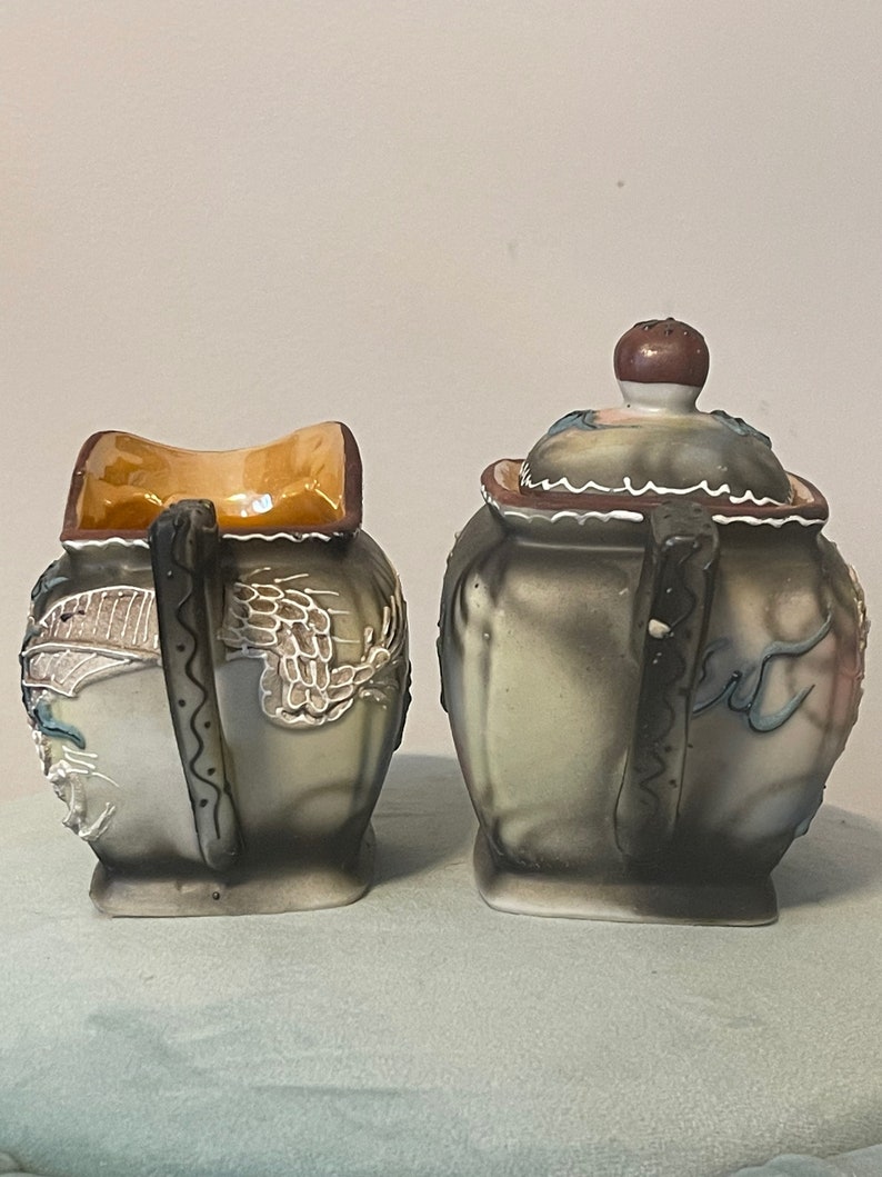 Op de afbeelding: Een vintage keramische suiker- en roomset met een drakenontwerp. De set is geschilderd in tinten grijs, wit, blauw en bruin. De roomkan heeft een bruin deksel en een gegolfde rand.