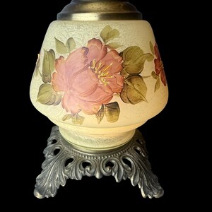 Puede incluir: Una l&aacute;mpara de mesa vintage con un dise&ntilde;o floral. La l&aacute;mpara presenta una pantalla en forma de campana de color crema adornada con una gran flor rosa y hojas verdes. Se apoya sobre una base de metal plateado ornamentada.