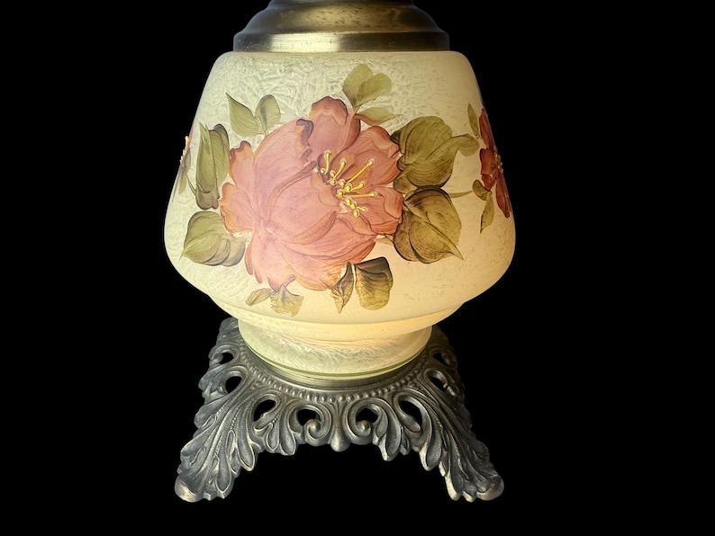 Puede incluir: Una l&aacute;mpara de mesa vintage con un dise&ntilde;o floral. La l&aacute;mpara presenta una pantalla en forma de campana de color crema adornada con una gran flor rosa y hojas verdes. Se apoya sobre una base de metal plateado ornamentada.
