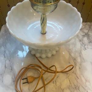 May include: A white milk glass lamp with a gold base and a white bowl-shaped shade. The lamp has a brown cord and plug.