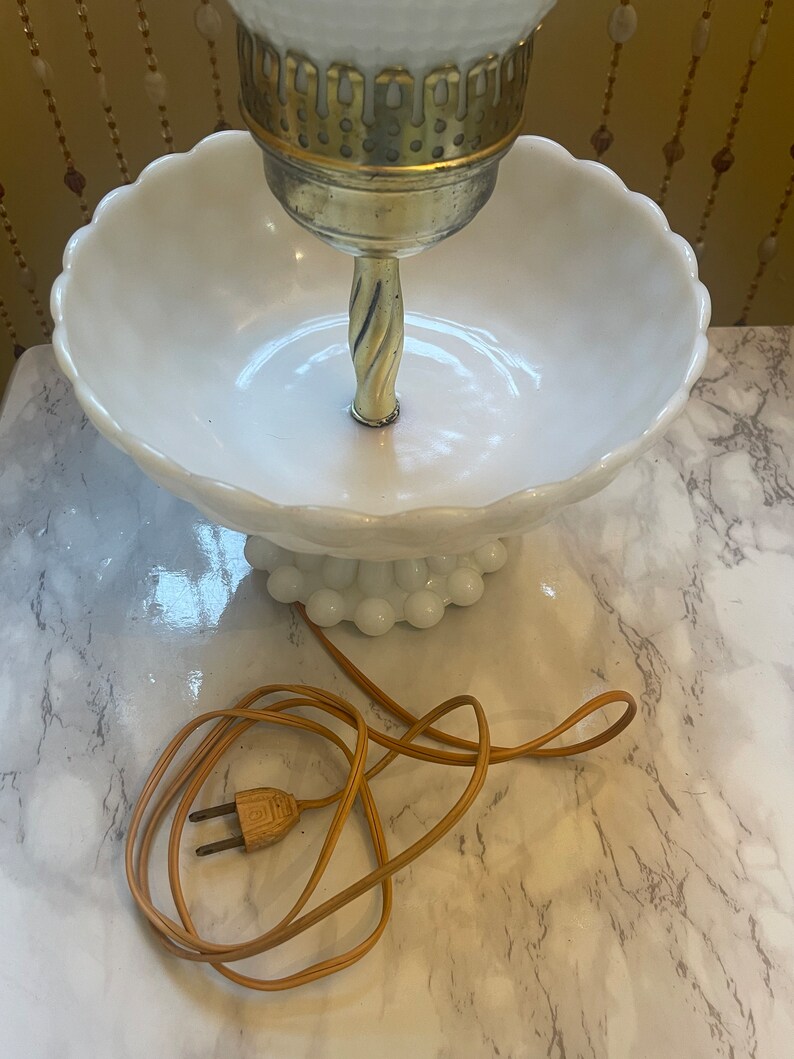 May include: A white milk glass lamp with a gold base and a white bowl-shaped shade. The lamp has a brown cord and plug.