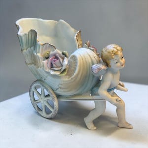 May include: A porcelain cherub figurine pushing a shell-shaped cart. The cart is decorated with pink roses and gold trim. The cherub has blonde hair and small wings. The cart has a single wheel.