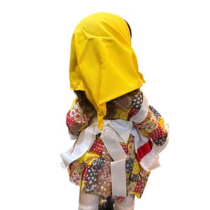 May include: A vintage doll with dark skin, wearing a yellow head covering and a patchwork dress in red, yellow, and brown. The doll has white socks and black shoes. White ribbons are tied around the waist.
