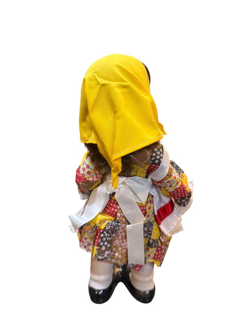 May include: A vintage doll with dark skin, wearing a yellow head covering and a patchwork dress in red, yellow, and brown. The doll has white socks and black shoes. White ribbons are tied around the waist.