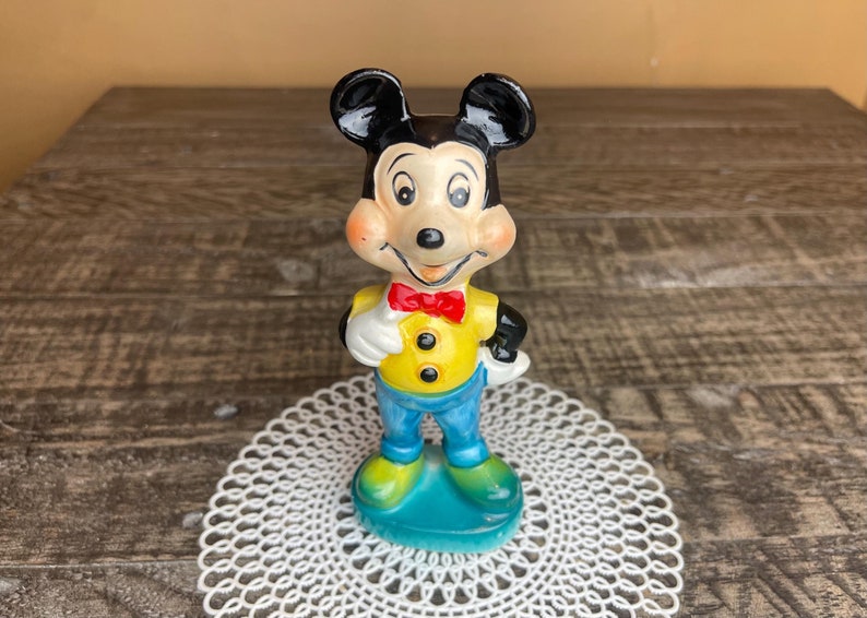 May include: A vintage ceramic figurine of Mickey Mouse wearing a yellow shirt, blue pants, and a red bow tie. He is standing on a blue base.
