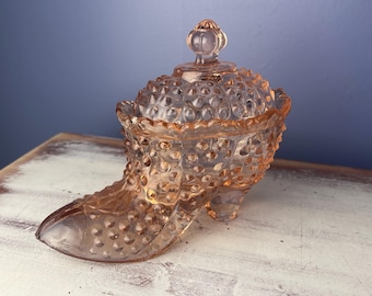 May include: A vintage, pink glass shoe-shaped trinket box with a hobnail pattern. The lid is topped with a small, round finial. The glass has a translucent quality, and the box sits on a white and brown surface.