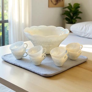 May include: A white ceramic serving bowl with a scalloped rim and grape vine design, surrounded by six matching teacups. The teacups and bowl are arranged on a light blue placemat, set on a wooden table.
