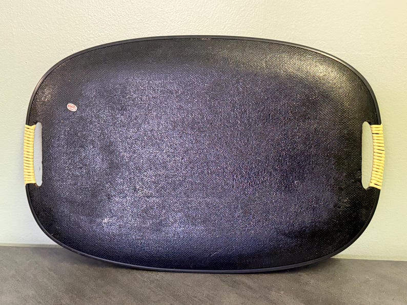 May include: Black oval serving tray with woven yellow handles. The tray has a textured surface and a dark color. The handles are made of a woven material. A small sticker is on the tray.