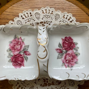 May include: A white porcelain serving dish with two sections and a handle. The dish is decorated with gold trim and pink roses.