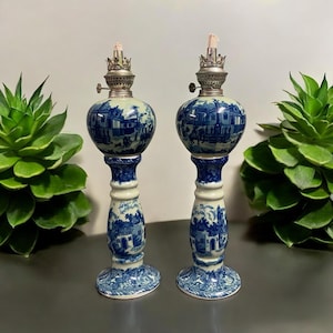 May include: Pair of blue and white ceramic oil lamps with a silver top and a floral design. The lamps are on a dark surface with green plants in the background.