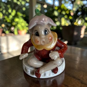May include: A ceramic figurine of a seated character wearing a red outfit with a yellow collar and a light purple flower-shaped hat. The figure has a pale face with rosy cheeks and is positioned on a white, circular, textured base with a scalloped edge.