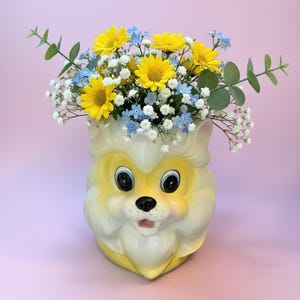 May include: A ceramic vase shaped like a stylized yellow and white animal head, filled with a bouquet of bright yellow daisies, small light blue forget-me-not flowers, white baby's breath, and green eucalyptus leaves with rounded foliage.