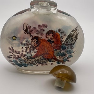 May include: A clear glass bottle with a painted design of two monkeys in a tree. The bottle has a brown mushroom-shaped stopper.
