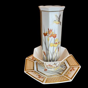 May include: A white ceramic vase with a geometric design and floral pattern featuring yellow and orange irises and hummingbirds. The vase is sitting on a white ceramic plate with a gold geometric pattern.