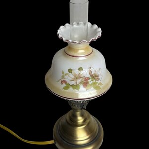 May include: A vintage-style table lamp with a floral design on the shade. The lamp features a brass base and a clear glass chimney. The shade is white with a yellow border and a floral pattern in pink and green.