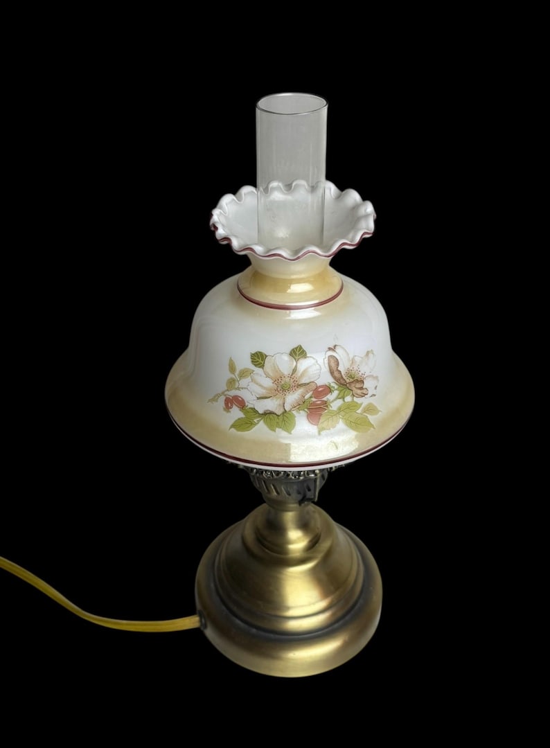 May include: A vintage-style table lamp with a floral design on the shade. The lamp features a brass base and a clear glass chimney. The shade is white with a yellow border and a floral pattern in pink and green.