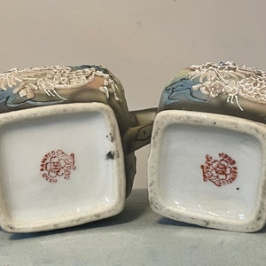 Op de afbeelding: Twee theepotten van keramiek met draken- en bloemenmotieven. De theepotten zijn geschilderd in tinten bruin, blauw en wit. De bodem van elke theepot heeft een rode stempel met de tekst "Hand Painted Japan".