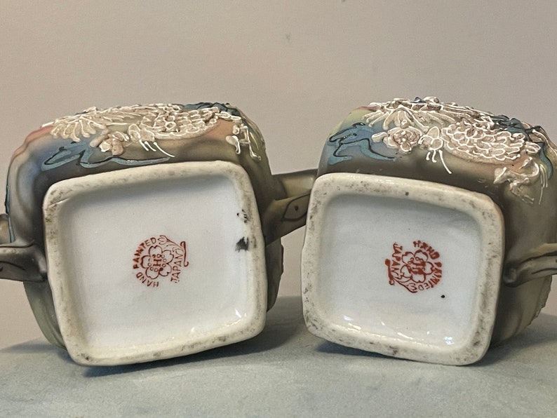 Op de afbeelding: Twee theepotten van keramiek met draken- en bloemenmotieven. De theepotten zijn geschilderd in tinten bruin, blauw en wit. De bodem van elke theepot heeft een rode stempel met de tekst "Hand Painted Japan".