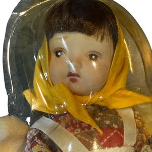 May include: A vintage doll with a painted face, dark hair, and a yellow headscarf. The doll is wearing a floral patterned dress with a white apron. The doll is encased in clear plastic.