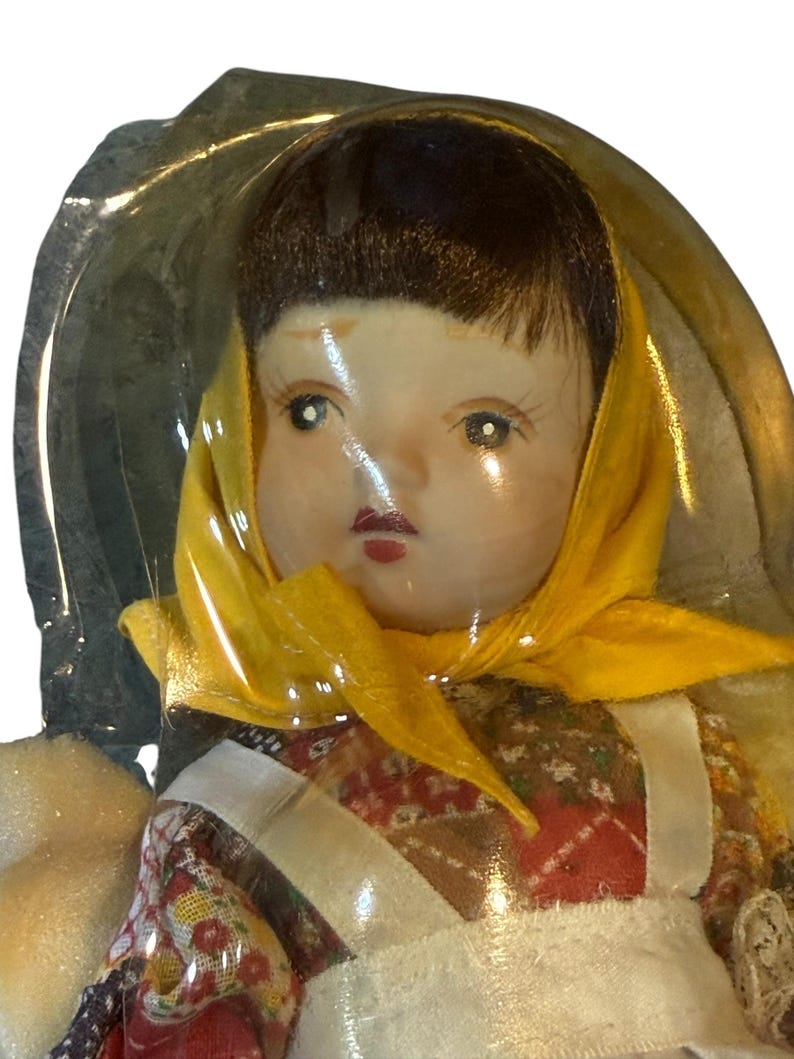 May include: A vintage doll with a painted face, dark hair, and a yellow headscarf. The doll is wearing a floral patterned dress with a white apron. The doll is encased in clear plastic.