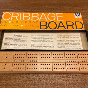 May include: A vintage Cribbage board game with a wooden pegboard and a box. The box is orange and white with the words "Cribbage Board" in large letters. The pegboard has multiple rows of holes for scoring. The game is designed for two to four players.