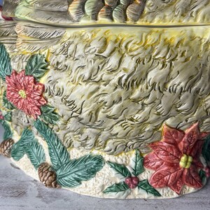 May include: A ceramic Christmas serving bowl shaped like a bird with a candy cane in its beak. The bowl is decorated with poinsettia flowers, holly, and pine branches.