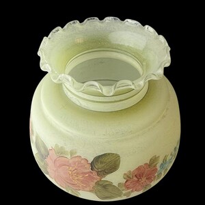 Puede incluir: Una pantalla de l&aacute;mpara de vidrio verde p&aacute;lido vintage con un borde con volantes. La pantalla de la l&aacute;mpara presenta dise&ntilde;os florales pintados a mano en tonos de rosa, rojo y azul, con hojas verdes. El vidrio tiene una apariencia texturizada y el fondo es negro.