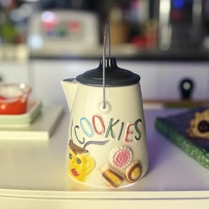 May include: Cream-colored ceramic cookie jar with a black lid and a metal handle. The word "COOKIES" is written in colorful letters. A cartoon image of a yellow face with a hat and cookies are also on the jar.