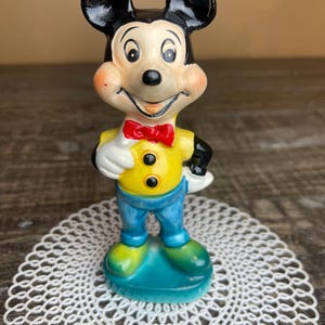 May include: A vintage ceramic figurine of Mickey Mouse wearing a yellow shirt, blue pants, and a red bow tie. The figurine is standing on a blue base.