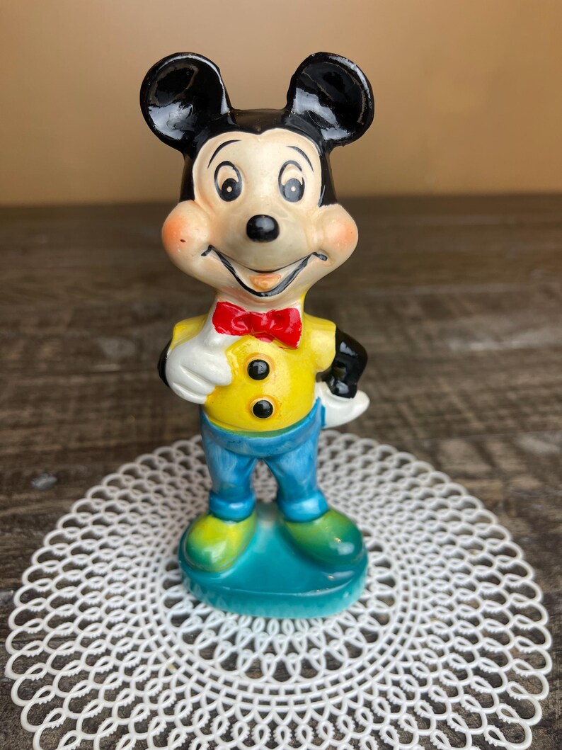 May include: A vintage ceramic figurine of Mickey Mouse wearing a yellow shirt, blue pants, and a red bow tie. The figurine is standing on a blue base.