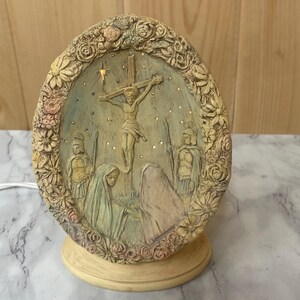 May include: A beige ceramic oval-shaped nightlight with a raised floral design. The center of the nightlight features a depiction of Jesus on the cross with a star above him. There are two figures on either side of the cross.