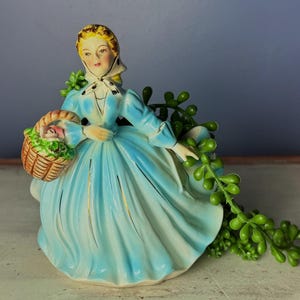 May include: A vintage porcelain figurine of a woman in a long, light blue dress, holding a woven basket filled with flowers. The figurine has gold accents and a white bonnet. Green foliage is behind the figurine.