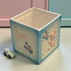 May include: A square, ceramic baby block planter with a light blue border. The block features a raised letter "A" and a pink teddy bear. Floral accents in blue and pink decorate the sides. A small white toy car sits nearby.