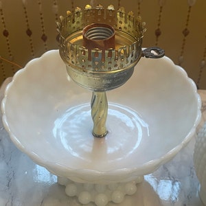 May include: A white milk glass pedestal with a gold metal lamp base. The lamp base has a decorative design and a light bulb socket.