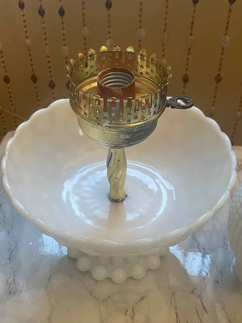 May include: A white milk glass pedestal with a gold metal lamp base. The lamp base has a decorative design and a light bulb socket.