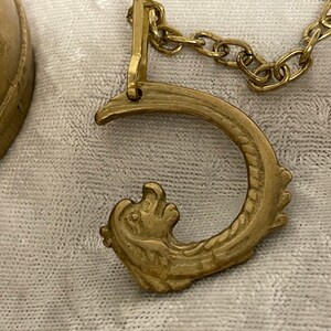 May include: A gold-colored metal bell with a decorative dragon-shaped handle. The bell is attached to a chain.
