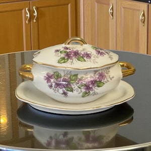 May include: A white porcelain soup tureen with a lid, handles, and a matching saucer. The tureen and lid are decorated with purple floral patterns and gold trim. The tureen is sitting on a black surface.
