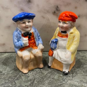 May include: Two vintage porcelain figurines of men wearing hats and sitting on stools. One man is wearing a blue hat and a blue jacket, the other is wearing a red hat and a yellow jacket.