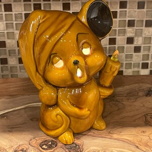May include: A ceramic mouse nightlight with a brown and black color scheme. The mouse is wearing a brown sleep mask and has a light bulb in its hand.
