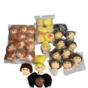 May include: A collection of doll heads with yarn hair in various colors including red, yellow, brown, and black. Some heads are packaged in clear plastic bags, while others are loose. Each head features painted facial details and soft vinyl skin.