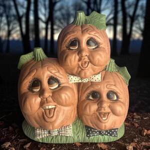 May include: A ceramic Halloween decoration featuring three smiling pumpkins with detailed faces, each wearing a bow tie. The pumpkins are in shades of orange and tan, with green leaves and a green base. The background is a blurred forest.