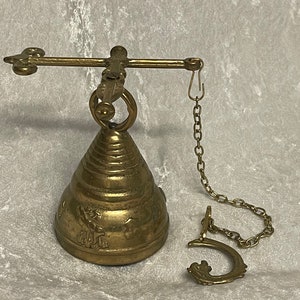 May include: A brass bell with a decorative design hangs from a chain attached to a metal bar. The bell has a conical shape and is adorned with intricate details.