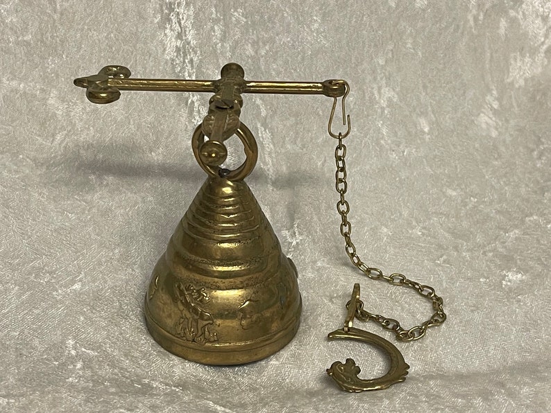 May include: A brass bell with a decorative design hangs from a chain attached to a metal bar. The bell has a conical shape and is adorned with intricate details.