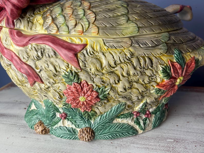 May include: A ceramic goose serving dish with a detailed, textured design. The goose is decorated with a red ribbon, green leaves, and red poinsettia flowers.