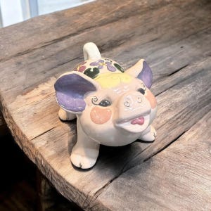 May include: A ceramic pig figurine with a cheerful expression. The pig has a light pink body, purple ears, and a floral design on its back. Its snout is pink, and its tongue is visible. The figurine is on a wooden surface.