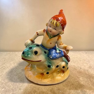 May include: A ceramic figurine depicting a small figure wearing a red pointed hat, a tan shirt, and blue pants, sitting atop a green and yellow spotted frog. The figure has blonde hair and is positioned with one hand resting on the frog's back.