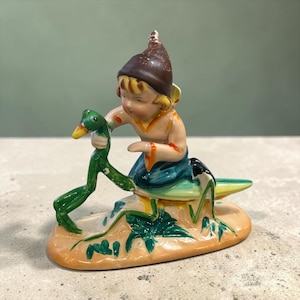 May include: A glazed ceramic figurine depicting a small figure wearing a brown pointed hat and orange-trimmed blue clothing, riding a large green grasshopper. The figure holds the grasshopper's head, and the entire piece rests on a tan base.