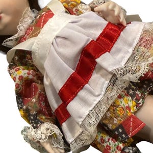 May include: A porcelain doll wearing a patchwork dress with a white apron and red ribbon accents. The dress features floral and geometric patterns, with lace trim on the sleeves and apron. The doll has brown hair and a serene expression.