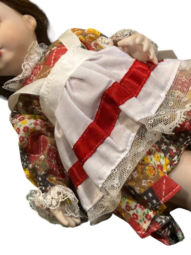 May include: A porcelain doll wearing a patchwork dress with a white apron and red ribbon accents. The dress features floral and geometric patterns, with lace trim on the sleeves and apron. The doll has brown hair and a serene expression.